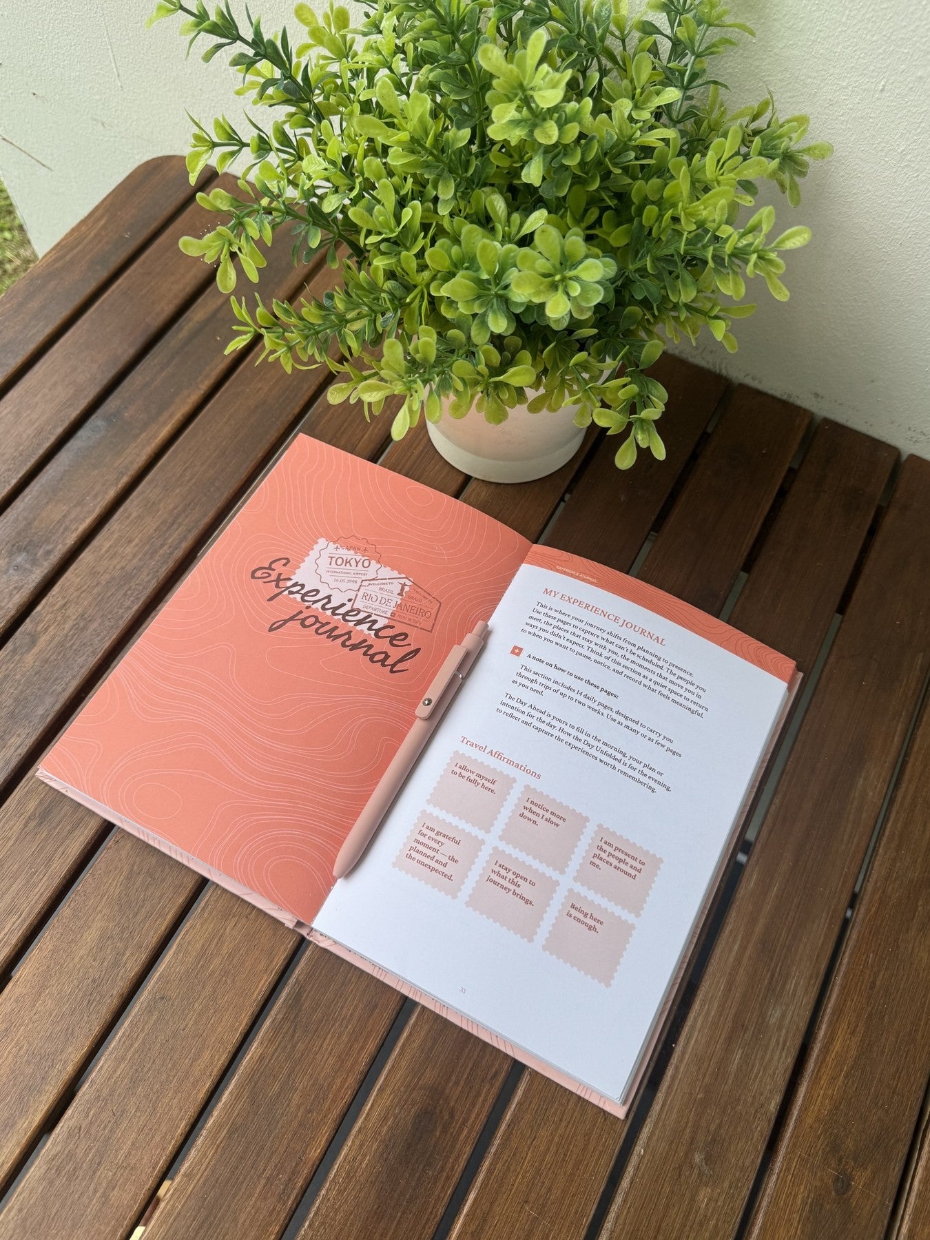 The coral Experience Journal beside an open journal page with daily prompts and travel affirmations, on a wooden table.