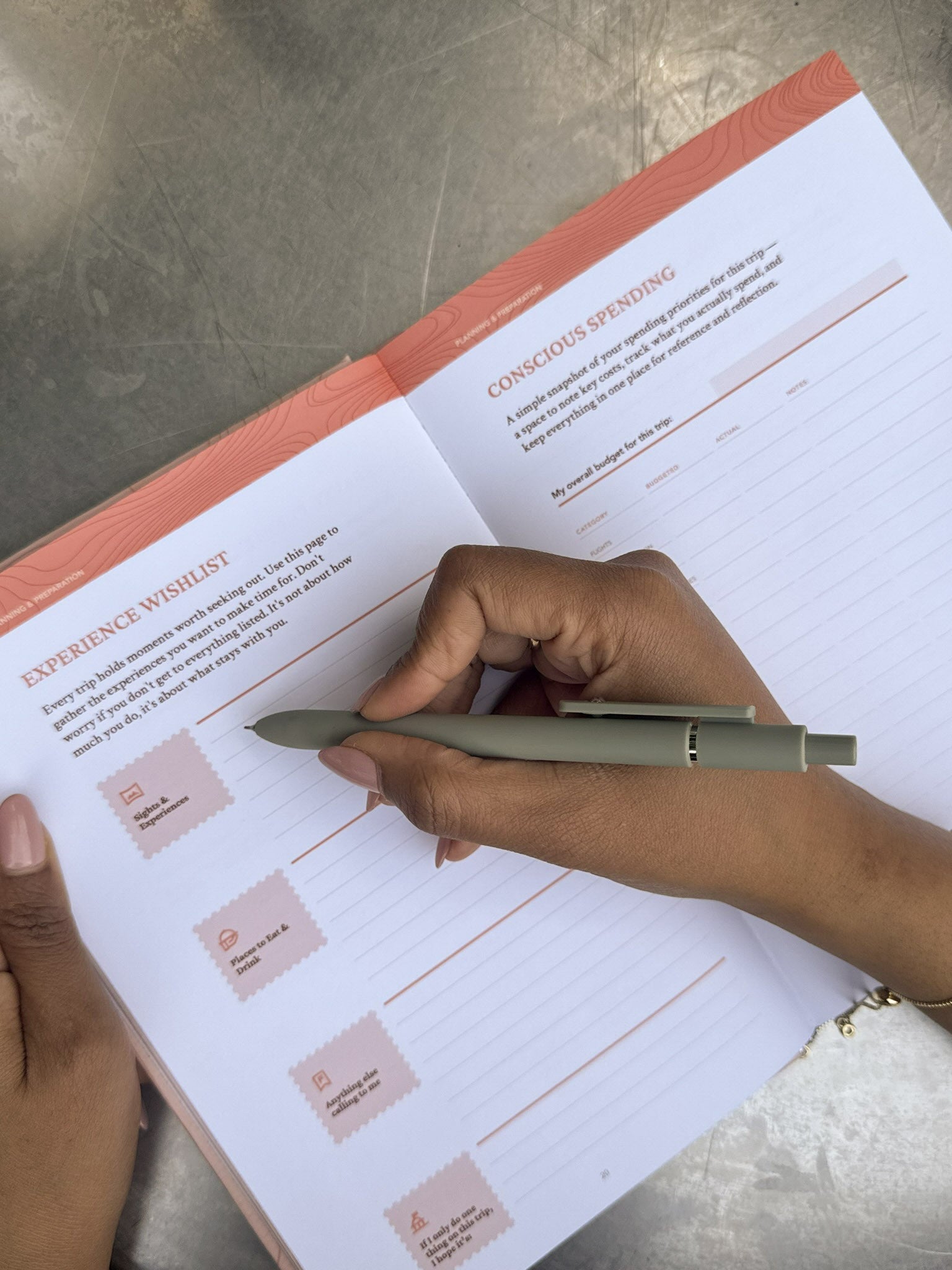 An open travel journal showing the Experience Wishlist and Conscious Spending pages as someone writes in the journal.